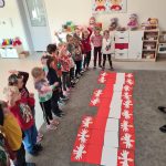 Dzieci stoją na szarym dywanie na wprost ułożonej dużej flagi Polski i ozdobionej godłami Polski. Podnoszą ręce do góry.