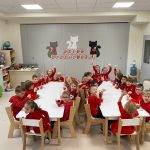 Dzieci w czerwonych bluzach siedzą w dwóch rzędach przy białych stołach, podnoszą ręce i machają.