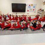 Dzieci ubrane na czerwono siedzą w dwóch rzędach. Podnoszą ręce do góry i wykonują ruchy do muzyki.