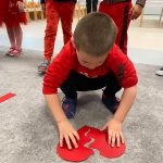 Chłopiec w czerwonej bluzie kuca na szarym dywanie i składa z dwóch części czerwone serce.