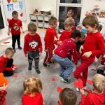 Bardzo dużo dzieci w czerwonych bluzach jest na szarym dywanie i szukają czerwone serca aby je złożyć i podarować.