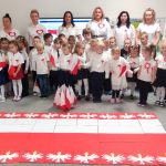 Szary dywan na nim ułożona jest duża flaga Polski otoczona białymi orłami. Naokoło niego stoi bardzo dużo dzieci, także dzieci niepełnosprawne oraz nauczyciele i dyrektor Przedszkola nr 12. Wszyscy ubrani odświętnie i śpiewają hymn ,,Mazurek Dąbrowskiego.