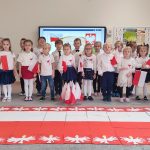 !9 dzieci stoi w dwóch rzędach na tle symboli narodowych. Dzieci stojące na przodzie trzymają flagi Polski. Przed nimi stoją flagi Polski oraz na dywanie ułożona jest duża flaga Polski otoczona białymi orłami.
