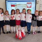 12 dziewczynek w strojach biało granatowych, biało czarnych i biało czerwonych stoi obok siebie trzymając się za ramiona. Przed nimi stoją małe flagi Polski, za nimi na tablicy symbole narodowe Polski.