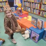 Dziewczynka w brązowym futerku stoi przed kolorowymi pufami na których stoją książki o misiach. O pufy oparte są pluszowe misie i książki.