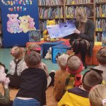 Na kolorowych pufach siedzi Pani w szarym swetrze, za plecami ma książki. W rękach trzyma książkę o niedźwiedziu którą czyta dzieciom, które siedzą przed nią.
