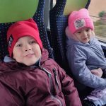 Chłopiec ubrany w kurtkę i czerwoną czapkę siedzi obok dziewczynki w niebieskiej kurtce i różowej czapce. Dzieci siedzą na fotelach w autobusie.