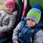 Chłopiec ubrany w kurtkę i szaro-zieloną czapkę siedzi obok dziewczynki w szarek kurtce i różowej czapce. Dzieci siedzą na fotelach w autobusie.
