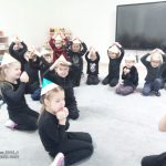 Dzieci w czarnych strojach, w czapkach w kształcie kredek siedzą na kolanach na szarym dywanie. Tworzą z rąk trójkąty nad głową.