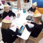 4 dzieci ubranych na czarno w czapkach w kształcie kredek, siedzą przy białym stole i rysują kredkami obrazek.