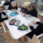 5 dzieci ubranych na czarno, w czapkach w kształcie kredek, siedzą przy białych stolikach i kolorują kredkami obrazek.