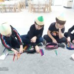4 chłopców ubranych na czarno w czapkach w kształcie kredki, siedzą na szarym dywanie i układają kredki wg. koloru i wielkości.