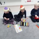 Troje dzieci ubranych na czarno siedzi na szarym dywanie. Na głowie mają czapki w kształcie kredek i układają kredki wg. wielkości.