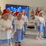 Dzieci ubrane w stroje biało-niebieskie stoją rozstawione na szarym dywanie i śpiewają piosenkę i kiwają rękami.