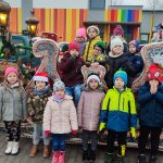14 dzieci ubranych w kurtkach i czapki, stoją przed saniami Św. Mikołaja zaprzężonymi w brązowego renifera.