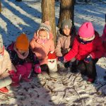 7 dziewczynek kuca w lesie na śniegu przy wizerunku małego Mikołaja. Dziewczynki są ciepło ubrane.