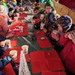 Dzieci ubrane w ciepłe, kolorowe kurtki siedzą na ławach za drewnianym stołem i spożywają smaczne pierniki i piją w kolorowych kubkach gorącą herbatę.