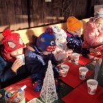6 dzieci ubrane w ciepłe, kolorowe kurtki siedzi na ławach za drewnianym stołem i spożywają smaczne pierniki i piją w kolorowych kubkach gorącą herbatę.