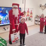 Chłopiec ubrany na czerwono w czapce Mikołaja stoi obok gwiazdora w czerwonym stroju i trzyma w ręce prezent: białe pudełko. Za nimi jest dekoracja świąteczna i jedno dziecko z paczką.