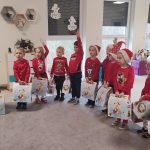 13 dzieci stoi w półkolu na szarym dywanie w rękach trzymają prezenty: białe kartony z wizerunkiem świątecznym. Niektóre dzieci się zgłaszają, aby porozmawiać z Gwiazdorem.