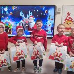 5 dzieci; chłopcy i dziewczynki ubrane na czerwono, w czapkach Mikołaja trzymają w ręce białe pudełka z prezentami, które otrzymały pod choinkę. Dzieci stoją na tle dekoracji świątecznej i choinki.
