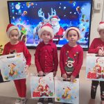 4 dzieci; chłopcy i dziewczynki ubrane na czerwono, w czapkach Mikołaja trzymają w ręce białe pudełka z prezentami, które otrzymały pod choinkę. Dzieci stoją na tle dekoracji świątecznej i choinki.