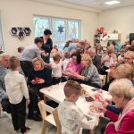 Zdjęcie przedstawia babcie i dziadków siedzących przy stołach i oglądających występy swoich wnuków, wszyscy są szczęśliwi i uśmiechnięci, sporo osób trzyma w ręku telefon i nagrywa.