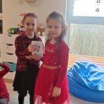 Stoją dwie dziewczynki ubrane na czerwono, stoją na szarym dywanie i wręczają sobie kartkę walentynkową.