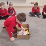 Chłopiec ubrany na czerwono klęczy na szarym dywanie przed szarym pudełkiem i wyciąga kartkę.