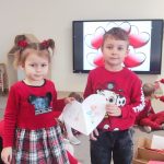 Stoi dziewczynka i chłopiec ubrani na czerwono, stoją na szarym dywanie i wręczają sobie kartkę walentynkową.