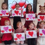 Dziewczynki ubrane na czerwono stoją obok siebie na tle szarej ściany i trzymają w rękach kartkę z czerwonymi sercami.