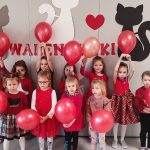 Dziewczynki ubrane na czerwono stoją obok siebie na tle szarej ściany i trzymają w rękach czerwone balony.