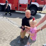 Dziewczynka z pomocą strażaka trzyma wąż i próbuje odkręcać wodę jak prawdziwy strażak.