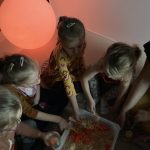 Zabawa sensoryczna. Dzieci siedzą w kółeczku. Za pomocą łyżeczek z plastikowego pojemnija, wkładają do słloiczków różnej wielkości pomarańczowe pomponiki.