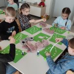 Dzieci siedzą przy stolikach i wykonują pracę plastyczną - zielony znaczek produktów ekologicznych.