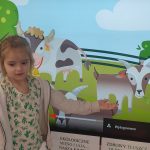Dziewczynka stoi na tle tablicy multimedialnej i pokazuje zwierzęta hodowlane.