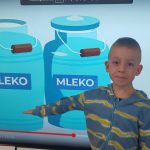 Chłopiec stoi na tle tablicy multimedialnej i pokazuje mleko.