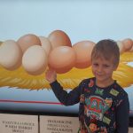 Chłopiec stoi na tle tablicy multimedialnej i pokazuje jajka.