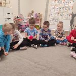 Dzieci siedzą na szarym dywanie obok siebie i trzymają czerwoną nitkę wełny.