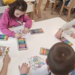 Dzieci siedzą przy białych stolikach i kolorują kolorowankę kredkami.