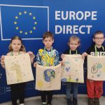 Dzieci na tle flagi Unii Europejskiej pozują do zdjęć z samodzielnie wykonaną torbą ekologiczną.