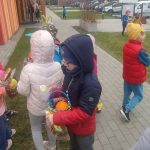 Dzieci wokół przedszkola na trawie szukają i znajdują koszyczki z prezentami.
