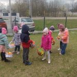 Dzieci wokół przedszkola na trawie szukają i znajdują koszyczki z prezentami.