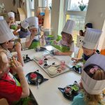 Dzieci w czapkach kucharskich siedzą przy białych stolikach i tworzą kolorowe desery z produktów ekologicznych.