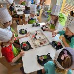 Dzieci w czapkach kucharskich siedzą przy białych stolikach i tworzą kolorowe desery z produktów ekologicznych.