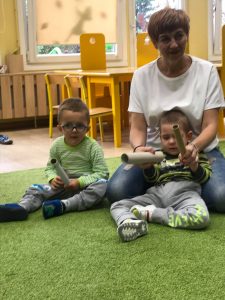 Dwoje dzieci siedzi na dywanie w tym jeden oparty jest o nauczycielkę w białej bluzce. Dzieci trzymają w rękach papierowe rurki od ręcznika papierowego,