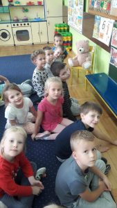 10 dzieci siedzi na dywanie i uśmiecha się do aparatu. Zvtyłu widać misia pluszowego posadzonego na krześle