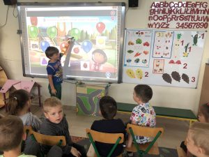 Chłopiec stojący przy tablicy interaktywnej rozwiązujący zadanie. Przed tablicą czworo dzieci siedzących na krzesłach.