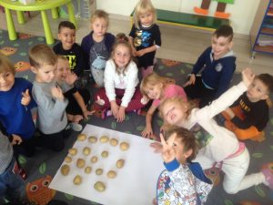 Grupa dzieci siedzących na dywanie i układających na białym brystolu wzór z ziemniaków.