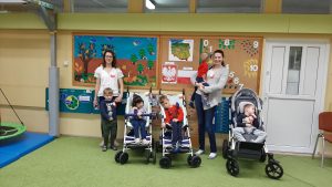 3 dzieci siedzi na swoim wózku. Po środku wózków stoi nauczycielka z dzieckiem na ręku. Po lewej stronie stoi nauczycielka trzymająca chłopca za rękę.. Każde dziecko ma przypięty biało-czerwony kotylion.